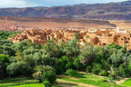 Tinghir to Merzouga scenic view showing a traditional brown mud-brick village surrounded by lush green palm trees and fields with mountains in the background