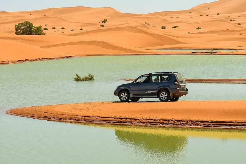Yakout Merzouga Luxury Camp featuring a black SUV parked on orange sand surrounded by a blue-green desert lake and dunes