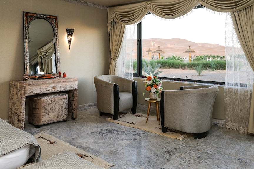 Luxury desert camp room at Yakout Merzouga Luxury Camp with beige walls, gray armchairs, decorative mirror, and large window view of sand dunes