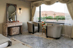 Luxury desert camp room at Yakout Merzouga Luxury Camp with beige walls, gray armchairs, decorative mirror, and large window view of sand dunes