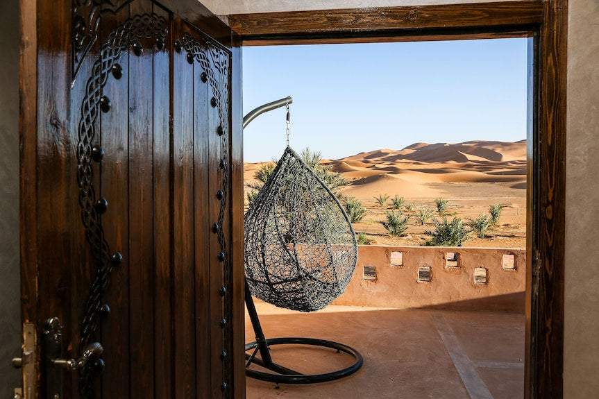 Yakout Merzouga Luxury Camp view of hanging black wicker chair on desert terrace with wooden door and Erg Chebbi dunes