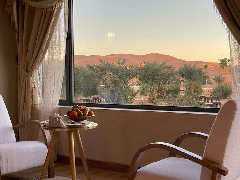 Interior view of Yakout Merzouga Luxury Camp featuring beige cushioned armchairs, a small wooden table with fruit, and large window facing palm trees and orange desert dunes