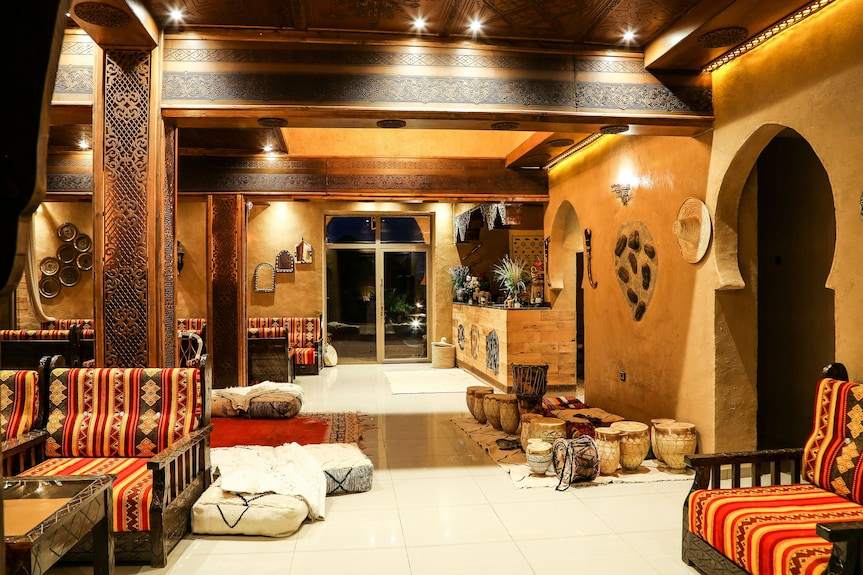 Yakout Merzouga Luxury Camp indoor lounge area featuring warm yellow walls, traditional Moroccan patterned cushions, wooden pillars, and tribal decor