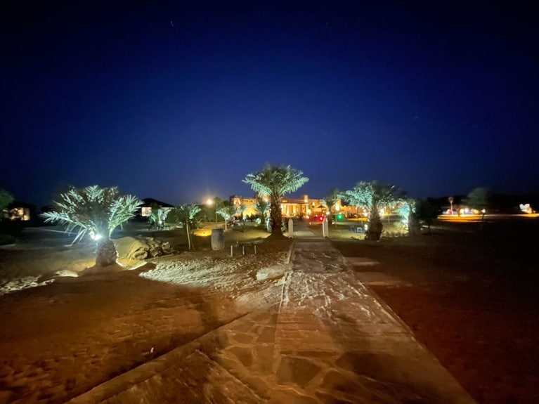 Yakout Merzouga Luxury Camp illuminated at night with palm trees and a stone pathway under a dark blue sky in the desert
