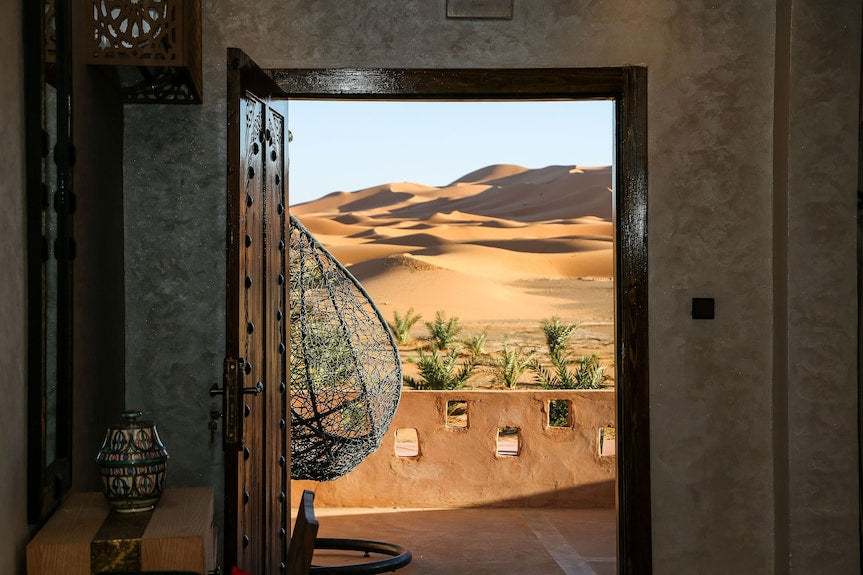 View of golden sand dunes through an open rustic wooden door with a hanging woven chair and desert wall at Yakout Merzouga Luxury Camp