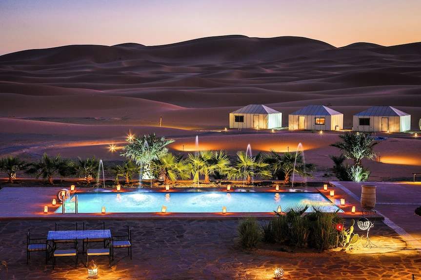 Yakout Merzouga Luxury Camp with illuminated white luxury tents near a glowing pool surrounded by desert dunes at sunset