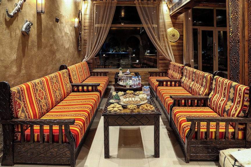 Yakout Merzouga Luxury Camp sitting area with traditional patterned orange cushions on dark wooden benches and a table with Moroccan snacks
