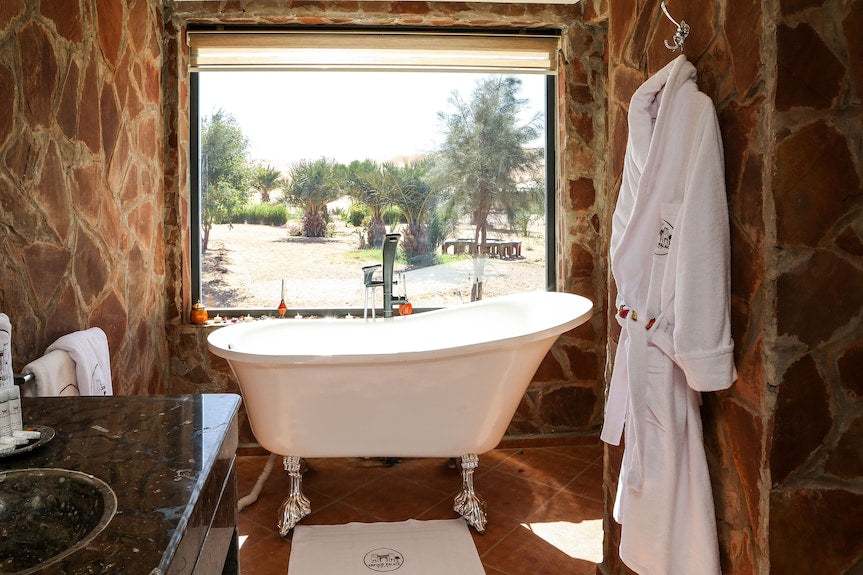 Luxury bathroom at Yakout Merzouga Luxury Camp featuring a white clawfoot bathtub, stone walls, large window view and hanging white bathrobe