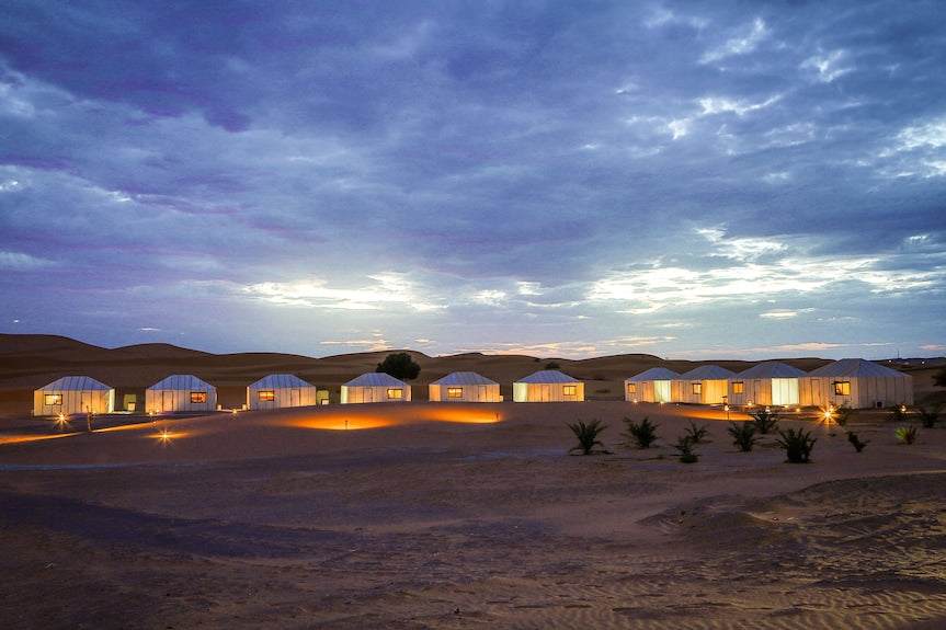 Yakout Merzouga Luxury Camp with illuminated white luxury tents in the sand desert under a cloudy twilight sky