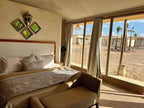 Yes We Camps Luxury Lodge & Spa bedroom with beige upholstery, patterned bedspread, decorative pillows, and large windows overlooking desert tents and palm trees