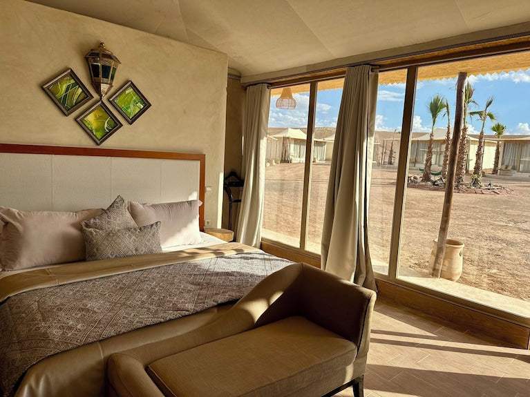 Yes We Camps Luxury Lodge & Spa bedroom with beige upholstery, patterned bedspread, decorative pillows, and large windows overlooking desert tents and palm trees