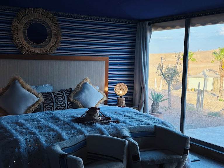 luxury bedroom inside Yes We Camps Luxury Lodge & Spa with blue striped walls, plush bedding, decorative cushions, and desert view window
