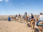 Camel caravan with tourists riding camels in colorful saddles crossing rocky desert terrain under blue sky at Yes We Camps Luxury Lodge & Spa