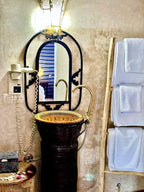 Bathroom interior featuring a decorative dark brown ceramic sink with gold accents, an oval mirror with black wrought iron frame, a wall-mounted hairdryer, and white towels on a wooden rack, showcasing the style of Yes We Camps Luxury Lodge & Spa