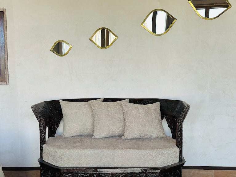 Dark carved wooden sofa with beige cushions and circular seat cushion near white wall with gold-framed eye-shaped mirrors