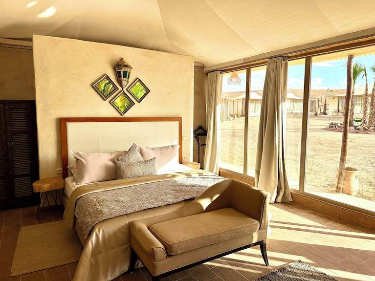 Yes We Camps Luxury Lodge & Spa bedroom with beige and brown bedding, cushioned bench, natural light, and desert view through large windows