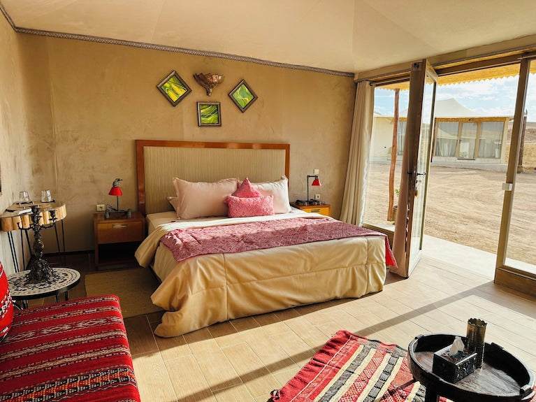 Luxury desert lodge bedroom with beige walls, double bed with pink and beige bedding, Moroccan-style red patterned seating, and large glass doors