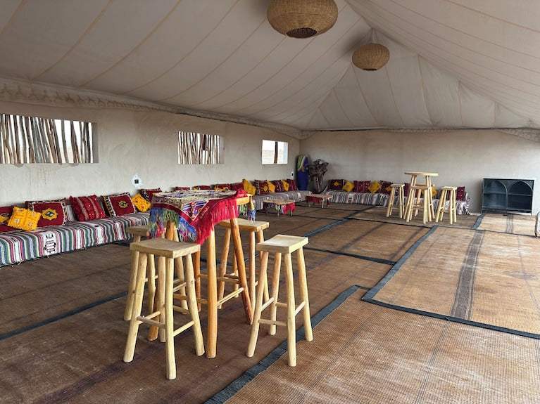 Interior of Yes We Camps Luxury Lodge & Spa featuring a spacious tent with woven floor mats, wooden stools, and colorful patterned cushions along low benches