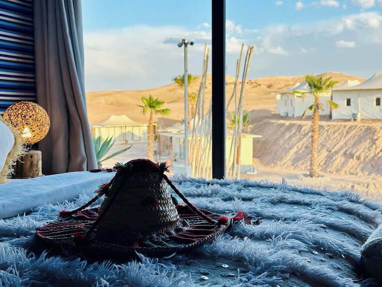 Yes We Camps Luxury Lodge & Spa room with a cozy bed, textured blanket, decorative woven hat, and desert landscape view