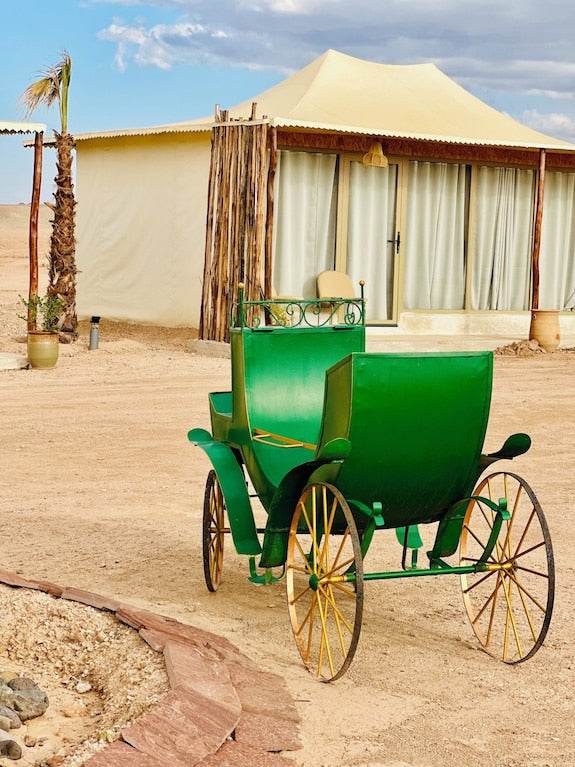 Green vintage horse-drawn carriage with yellow wheels in the desert in front of a beige luxury tent, showcasing Yes We Camps Luxury Lodge & Spa