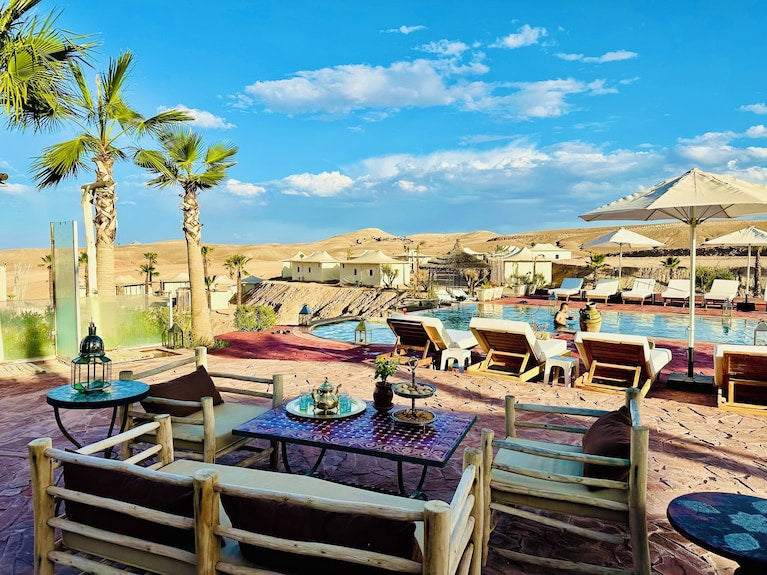 Yes We Camps Luxury Lodge & Spa outdoor seating area with wooden furniture, pool, umbrellas, and desert landscape under blue sky