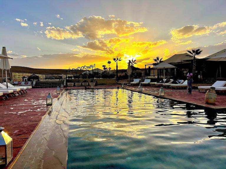 Outdoor swimming pool surrounded by lounge chairs and umbrellas at Yes We Camps Luxury Lodge & Spa with a vibrant sunset sky