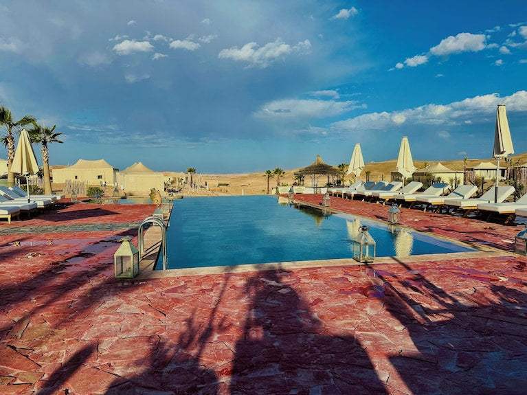 Outdoor rectangular swimming pool with stone red tile deck, surrounded by white lounge chairs and umbrellas at Yes We Camps Luxury Lodge & Spa