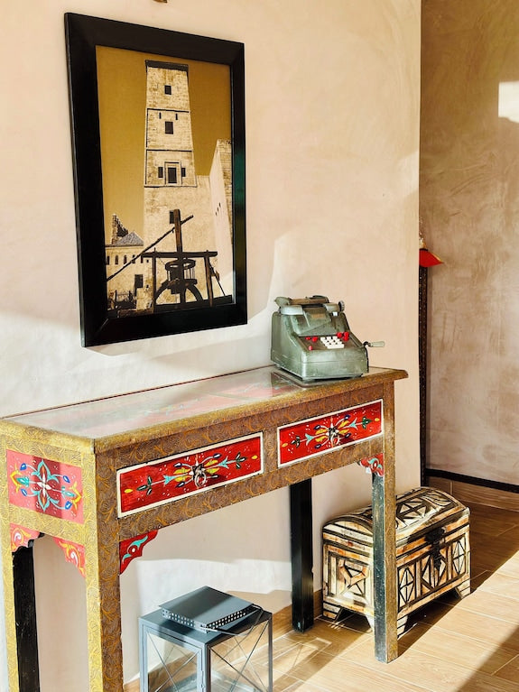 Decorative wooden desk with red floral painted drawers and a geometric patterned ottoman underneath, featuring a vintage cash register and framed tower art above