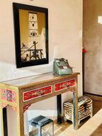 Decorative wooden desk with red floral painted drawers and a geometric patterned ottoman underneath, featuring a vintage cash register and framed tower art above