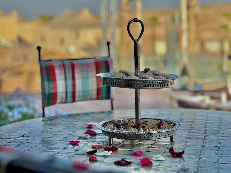 Yes We Camps Luxury Lodge & Spa metal two-tier silver serving tray with pastries on patterned tile table surrounded by plaid cushions and rose petals